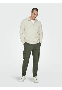 Beige Sweatshirt mit Viertelreißverschluss und glatter Textur, kombiniert mit olivfarbenen Cargo-Hosen mit zwei seitlichen Taschen. Das Modell trägt weiße Sportschuhe.