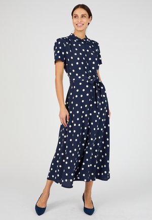 Mujer con un vestido midi azul marino con lunares blancos, mangas cortas, cuello, cinturón en la cintura y tacones azul marino, sonriendo y posando.