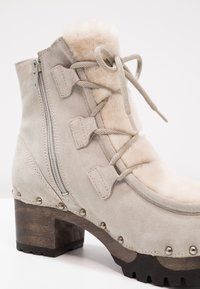 Bottine beige en daim avec doublure en fourrure, lacets sur le devant, fermeture éclair et talon bloc rond en bois. Présente des clous en métal le long de la semelle.