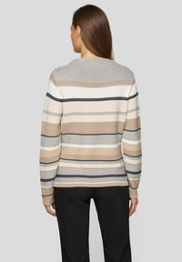 Frau trägt einen langärmligen, gestrickten Pullover mit horizontalen beigen, weißen, grauen und schwarzen Streifen, kombiniert mit schwarzen Hosen, mit dem Rücken zur Kamera.