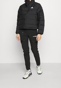 Giacca a vento nera con colletto alto, tasche con zip e logo Adidas; abbinata a pantaloni softshell neri con zip laterali e branding Terrex.