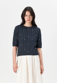 Navy blue short-sleeve cable knit sweater featuring decorative pearl accents, ribbed cuffs, and hem. Worn over a white skirt.