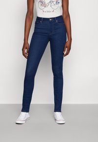 Mujer con jeans ajustados azul oscuro, top de tirantes blanco con gráfico floral y zapatillas blancas, de pie contra un fondo liso.