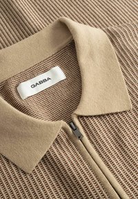 Beige ja pruuni värvi käsitsi kootud kampsuni kaeluse lähivõte, kus on tõmblukk ja valge silt kirjaga "GABBA".