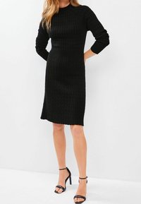 Robe en tricot côtelé noire avec un col rond et des manches longues, silhouette ajustée, ourlet au niveau du genou et motif texturé. Associée à des sandales noires.