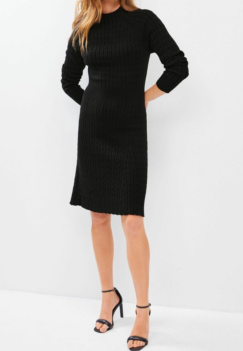 Robe en tricot côtelé noire avec un col rond et des manches longues, silhouette ajustée, ourlet au niveau du genou et motif texturé. Associée à des sandales noires.