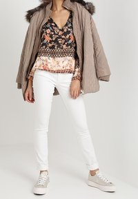 Femme portant une veste matelassée beige avec capuche doublée de fourrure sur un chemisier fleuri, pantalon skinny blanc et baskets beiges avec semelles blanches.