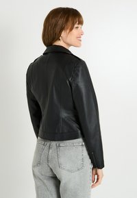 Femme portant une veste en cuir noire et un jeans gris clair, souriant et regardant sur le côté contre un fond blanc uni.