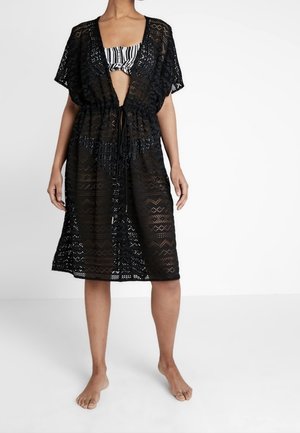 Kobieta ubrana w czarny, szydełkowany, sięgający do kolan okrycie na czarno-biały, paskowany top bikini, stojąca boso na jednolitym tle.