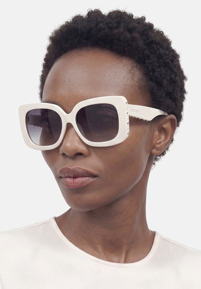 Femme aux cheveux courts et bouclés portant de grandes lunettes de soleil blanches à verres foncés et un haut blanc sur un fond uni.