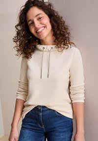 Beżowa bluza z kapturem o luźnym kroju, z ozdobnym ściągaczem przy szyi i prążkowanymi mankietami, w komplecie z ciemnoniebieskimi dżinsami.