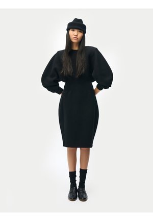 Frau trägt ein schwarzes Kleid mit Puffärmeln, schwarze Mütze, schwarze Socken und schwarze Loafer, steht mit den Händen in der Hüfte vor weißem Hintergrund.