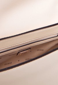 Interno di una borsa a mano color beige chiaro con una texture liscia, dotato di una tasca con zip e il nome del marchio "Guess" stampato ripetutamente sul rivestimento.