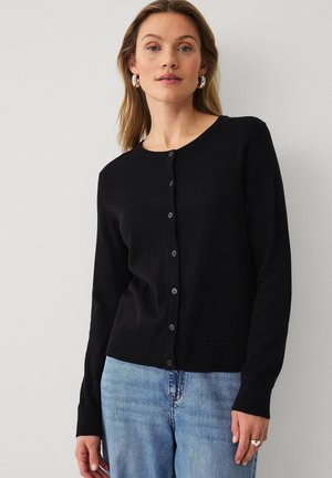 Frau trägt eine schwarze, geknöpfte Strickjacke und hellblaue Jeans, steht vor einem einfachen hellgrauen Hintergrund und blickt nach vorn.