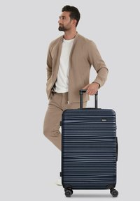 Hombre vestido con un traje beige, de pie con una mano en el bolsillo y la otra sosteniendo el mango de una gran maleta azul marino con ruedas.