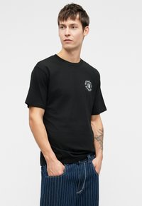 Zwarte katoenen T-shirt met korte mouwen, ronde halslijn, met een witte grafische print op de linkerkant van de borst. Gecombineerd met blauwe gestreepte spijkerbroeken.