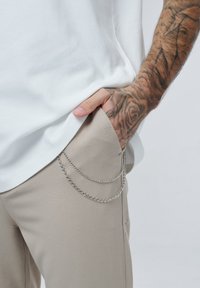 Pravá ruka s detailními tetováními v kapse béžových kalhot, na sobě má bílé tričko a stříbrné řetězy visící z kapsy kalhot.