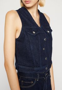 Ciemnoniebieska kamizelka z denimu z dekoltem w serek, bez rękawów, z dwoma kieszeniami na piersi i zapinana na guziki z przodu. Posiada krój skrócony i widoczne przeszycia.