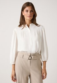 White button-up blouse with a stand collar, loose sleeves, and gathered detail; paired with high-waisted beige trousers featuring a belt.