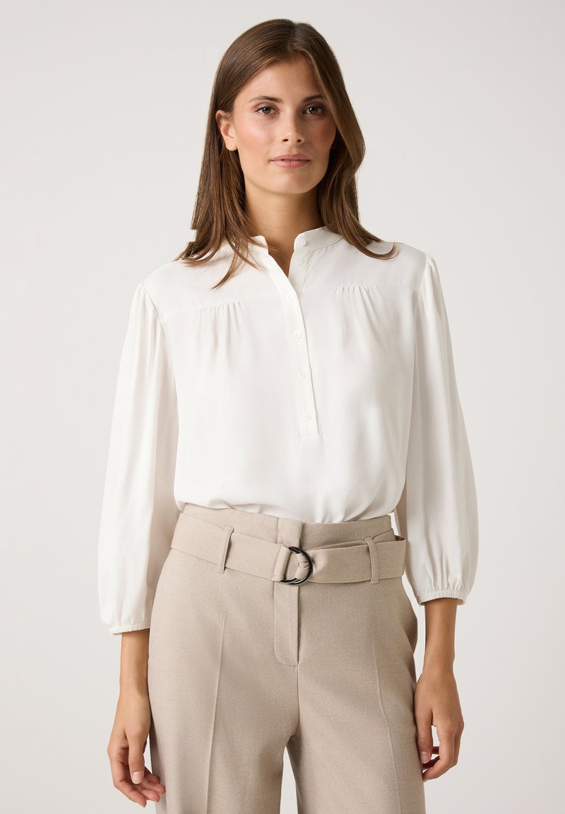 White button-up blouse with a stand collar, loose sleeves, and gathered detail; paired with high-waisted beige trousers featuring a belt.
