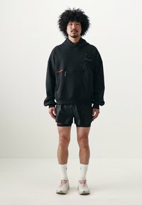 Sort hættejakke med røde detaljer, parret med sorte shorts. Fodtøjet er lysefarvet. Enkelt, funktionelt design med en afslappet pasform.