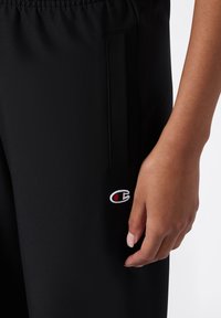Zwarte sportieve broek met een elastische tailleband, zijzak en een klein geborduurd logo in rood en wit nabij de zoom.