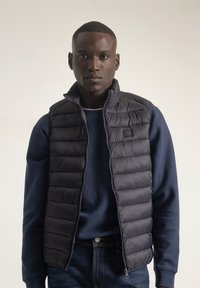 Chaleco acolchado en azul marino oscuro con diseño de acolchado y cuello alto, superpuesto sobre un sudadero azul navy. Presenta bolsillos con cremallera y un detalle de logo.