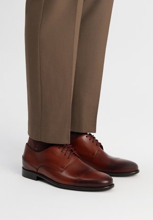 Braune Lederschuhe mit glatter Textur, abgerundetem Zeh und Schnürdesign. Kombiniert mit braunen Hosen und dunklen Socken.