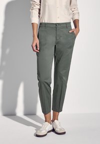 Zielone, dopasowane spodnie z guzikiem i bocznymi kieszeniami. Noszone z białymi trampkami. Gładka faktura, elegancki krój, krótsza długość.