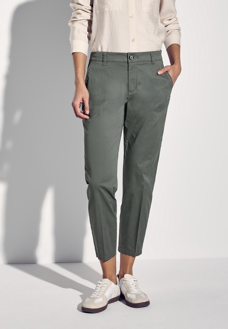 Zielone, dopasowane spodnie z guzikiem i bocznymi kieszeniami. Noszone z białymi trampkami. Gładka faktura, elegancki krój, krótsza długość.