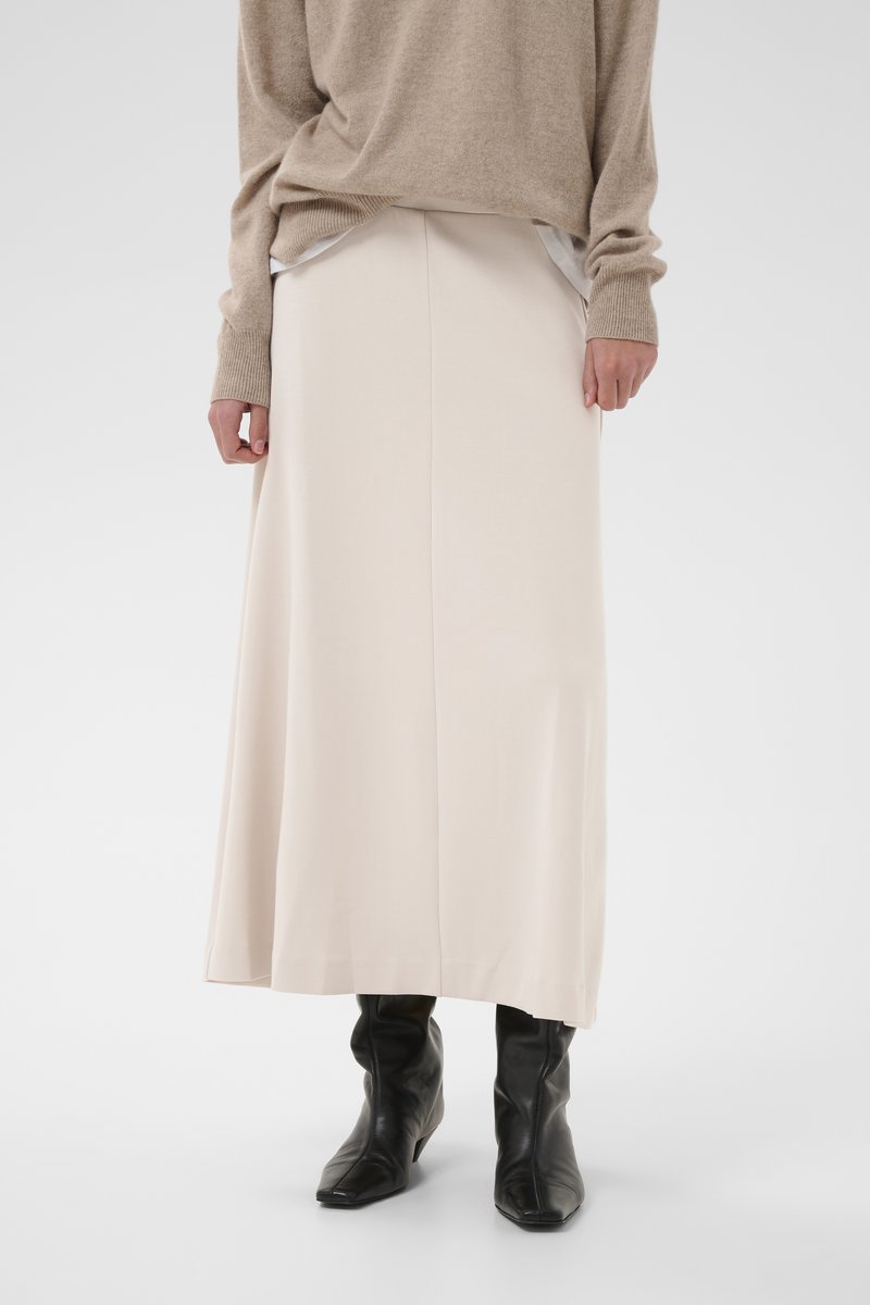 Beež pikk seelik, millel on sile kangapind, peene küljesõmbluse ja lõdva lõikega, kombineeritud teravate mustade pahkluu saapadega.