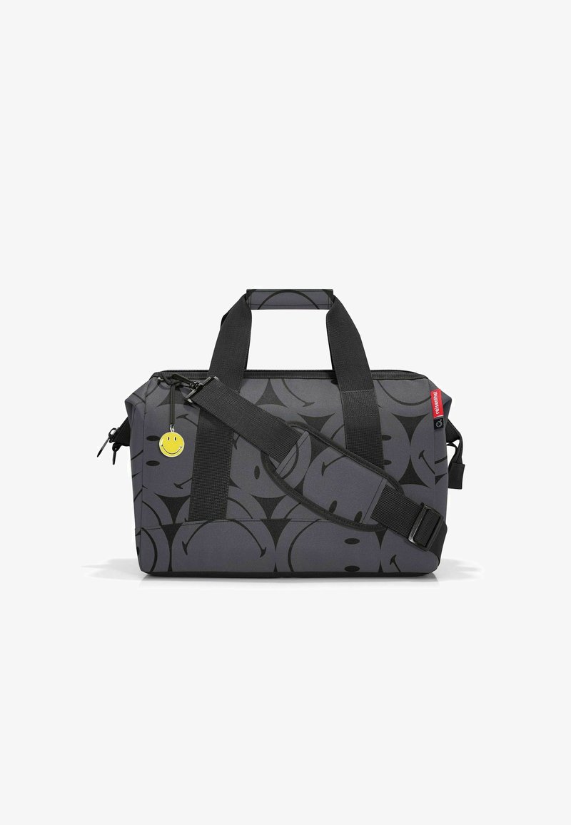 Sac de voyage en tissu gris avec un motif de smiley noir, poignées en sangle noires, bandoulière ajustable et un porte-clés smiley jaune.