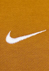 Nike-logo, witte geborduurde swoosh op een gestructureerde, goudbruin stoffen achtergrond. De stof heeft een ribbelpatroon en voelt glad aan.