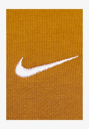 Nike-logo, witte geborduurde swoosh op een gestructureerde, goudbruin stoffen achtergrond. De stof heeft een ribbelpatroon en voelt glad aan.