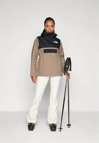 Žena v béžovo-čiernej lyžiarskej bunde, bielych lyžiarskych nohaviciach a čiernych lyžiarskych topánkach, drží dve čierne lyžiarske palice na jednofarebnom pozadí.