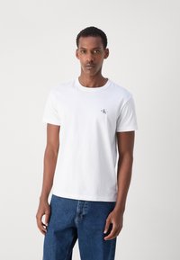 Hvid bomulds t-shirt med korte ærmer og rund halsudskæring, med et lille sort logo på venstre bryst. Sammen med blå denim jeans.
