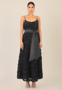 Vestido negro con flores que presenta aplicaciones de rosas 3D en capas, tirantes finos y un cinturón de lazo de satén en la cintura, combinado con zapatos de tacón plateados.