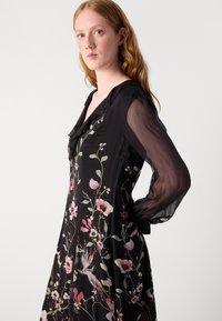 Schwarzes Kleid mit floralen Mustern in Pink und Weiß, transparenten langen Ärmeln, rüschigem Ausschnitt und fließender Silhouette.