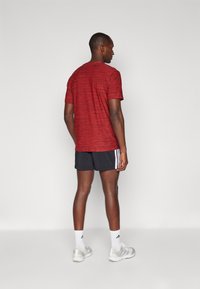 T-shirt de sport rouge à manches courtes avec un motif texturé, associé à un short noir avec des rayures blanches, porté avec des chaussettes blanches et des baskets.