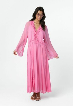 Žena v dlhej ružovej priesvitnej sukni s volánikovým výstrihom do V, zvonovými rukávmi a pásom na zaväzovanie, doplnená ružovými slnečnými okuliarmi a sandálmi.