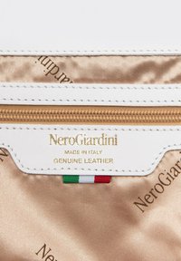 Vit läderpås med guldfärgad dragkedja, med ett logotypprytt innerfoder och en italiensk flagga som accent. Etiketten visar "NeroGiardini, Made in Italy, Äkta läder."
