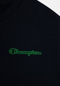 T-shirt nero in cotone con il logo "Champion" ricamato in verde sul petto. Il tessuto ha una consistenza liscia con un design classico a girocollo.