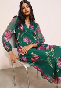 Kukkamainen mekko vihreästä kankaalta, jossa on vaaleanpunaisia tulppaanikuvioita. Sisältää läpikuultavat pitkät hihat ja V-pääntien. Malli istuu hopeisella tuolilla.