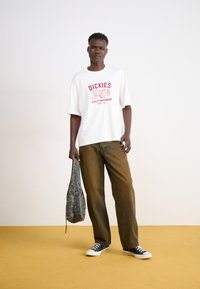 T-shirt bianco a maniche corte con testo grafico rosso, abbinato a pantaloni marroni larghi e sneaker neri. Tiene una borsa grigia a fantasia.