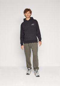 Felpa nera con tasca a marsupio anteriore e logo bianco, abbinata a pantaloni della tuta verde oliva e scarpe da ginnastica grigie con accenti.