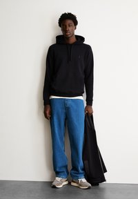 Felpa nera con cappuccio e tasca frontale, jeans blu larghi e scarpe da ginnastica grigie. Il modello tiene in mano una giacca nera, in piedi davanti a una parete chiara.