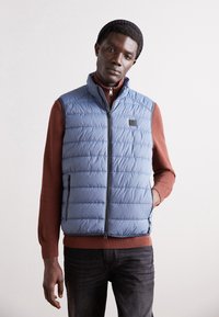 Světle modrá péřová vesta s vodorovným prošíváním, předním zipem a postranními kapsami, nošená přes rustikální svetr a tmavé džíny.