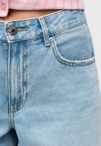 Nærbilde av livet på lyseblå denimjeans med knapp, beltehempe, frontlomme og en del av en rosa skjorte som er stukket inn over.