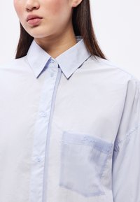 Svetlomodrá košeľa s golierom vyrobená z hladkej tkaniny, s gombíkmi a ľavým náprsným vreckom so vyšívaným logom.