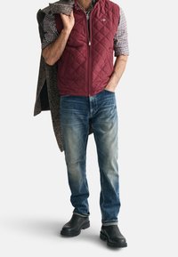 Bordeaux gewatteerd vest over een geruite shirt, blauwe jeans en zwarte leren enkellaarsjes, met een gelaagde textuur en casual ontwerp.